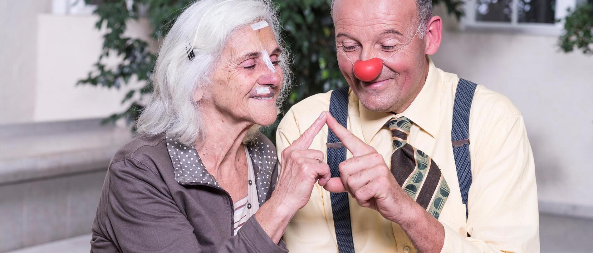 Ein Mann mit roter Clownsnase sitzt neben einer Seniorin; beide lächeln während sich ihre Fingerspitzen berühren.