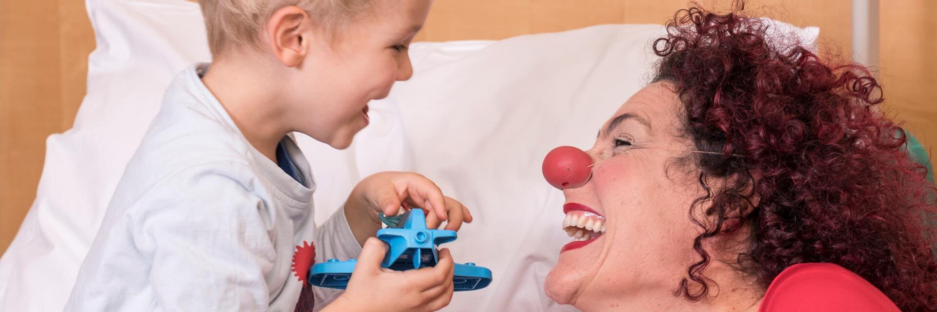 Ein Kind mit einem Spielzeug in der Hand sitzt in einem Bett, daneben sitzt eine Clownin; beide lachen herzhaft.
