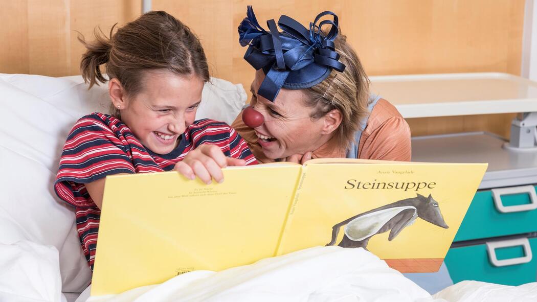 Eine Clownfrau schaut gemeinsam mit einem Mädchen ein Buch an; beide lachen.