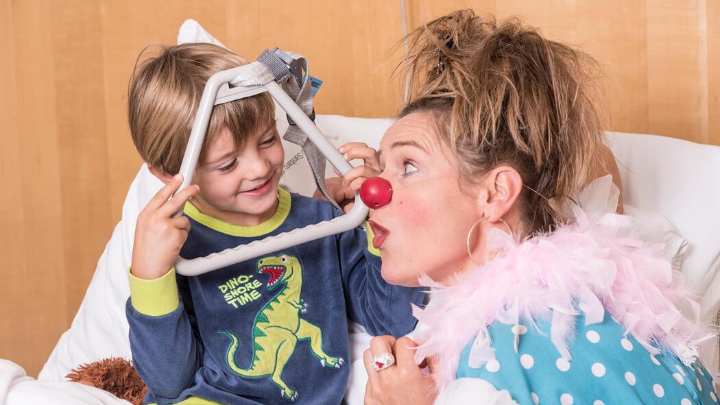 Ein Kind blickt lachend durch den Rahmen der Aufrichthilfe am Krankenbett, während eine Clownin mit roter Nase daneben überrascht schaut