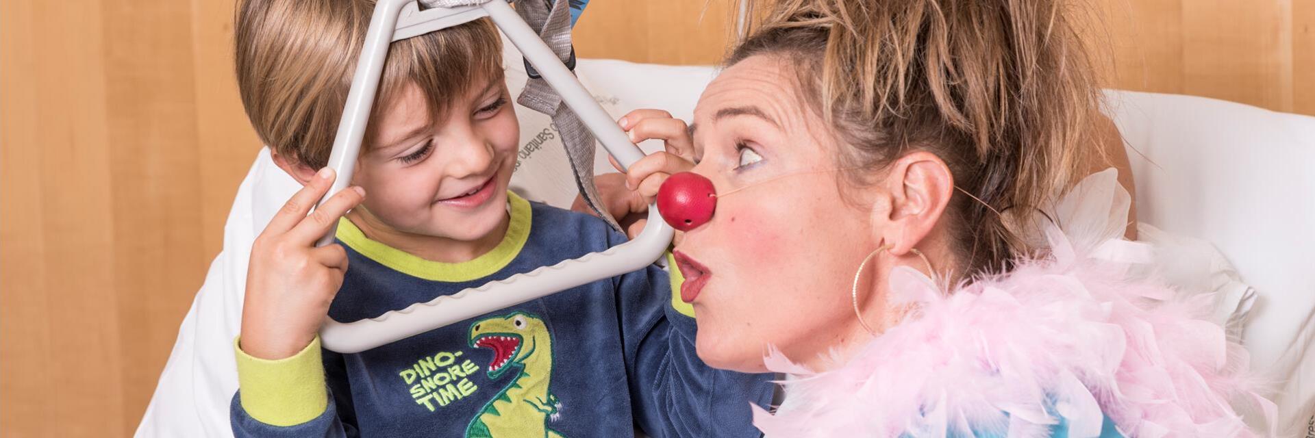 Ein Kind blickt lachend durch den Rahmen der Aufrichthilfe am Krankenbett, während eine Clownin mit roter Nase daneben überrascht schaut