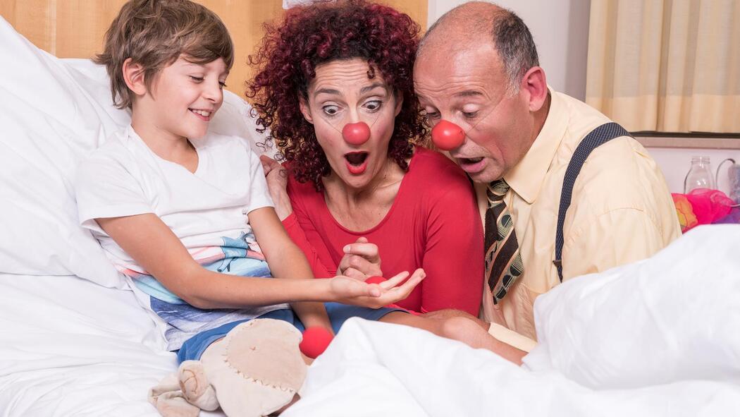 Zwei Clowns sitzen bei einem lachenden Kind am Bett und schauen erstaunt auf seine Hand.