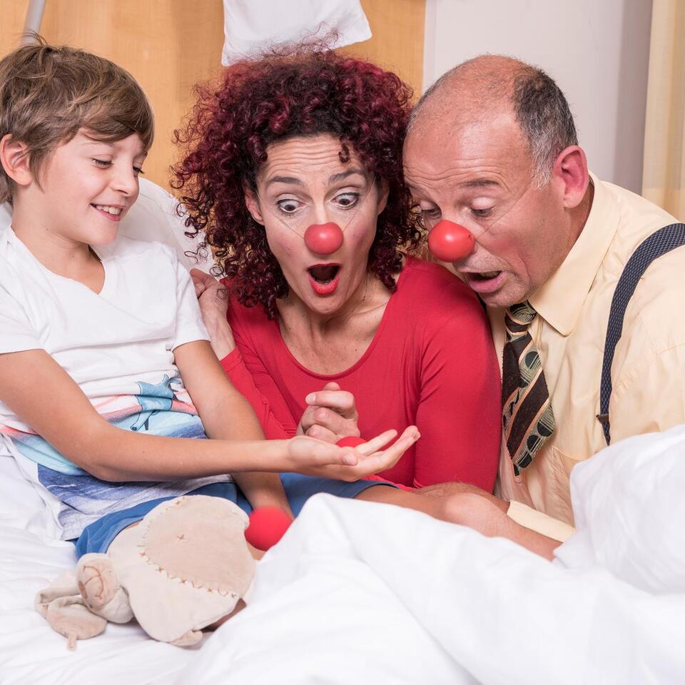 Zwei Clowns sitzen bei einem lachenden Kind am Bett und schauen erstaunt auf seine Hand.