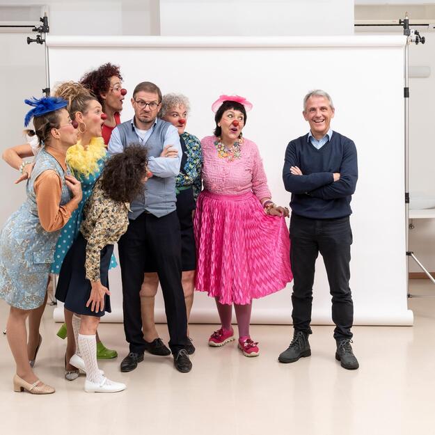 Mehrere Clowninnen mit roten Nasen und bunten Kleidern posieren gemeinsam mit zwei Männern vor einer weißen Leinwand.