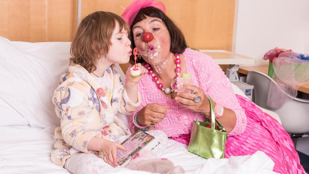 Ein kleines Mädchen sitzt auf einem Bett, daneben eine Clownfrau in einem Rosa Kleid; die beiden machen Seifenblasen.