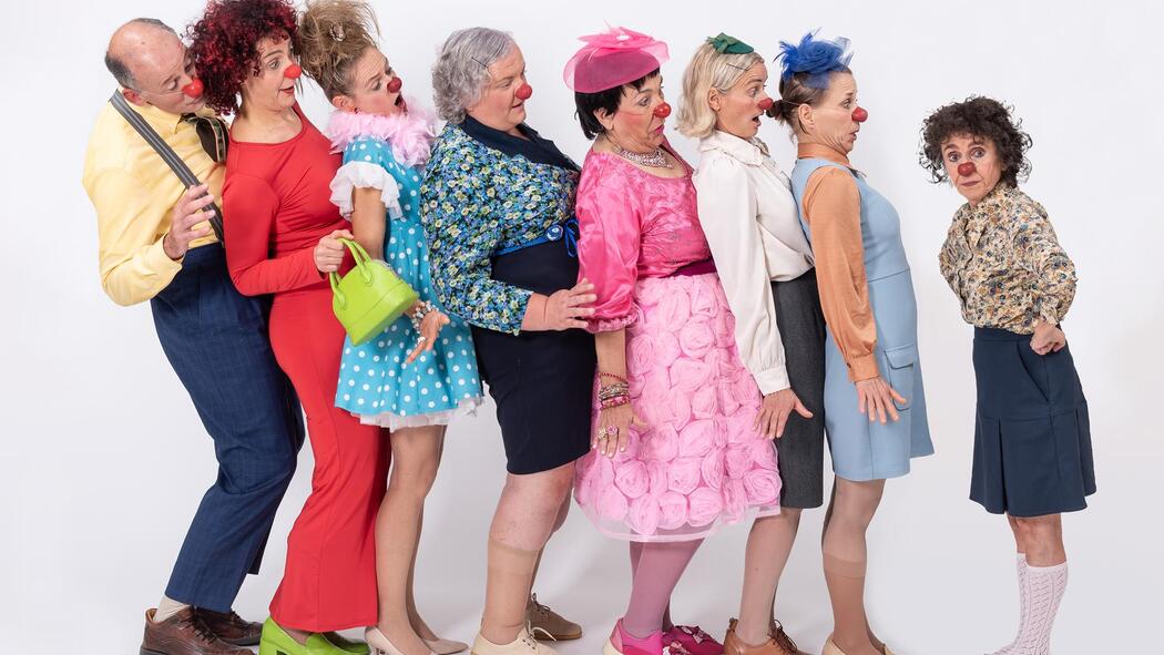 Mehrere Clowninnen mit roten Nasen und bunten Kostümen stehen dicht hintereinander und beugen sich nach hinten; ihnen gegenüber steht eine weitere kleinere Clownin.