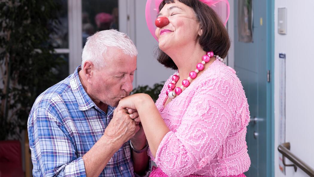 Eine Clownin in einem rosa Kleid und mit Hut freut sich über den Handkuss eines älteren Mannes in einem blau-weiß karierten Hemd.