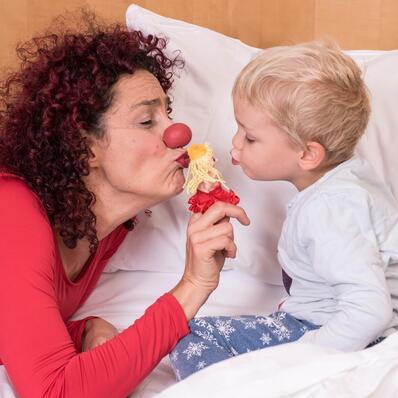 Eine Clownfrau unterhält mit einer kleinen Fingerpuppe ein kleines Kind, das in einem Bett sitzt.