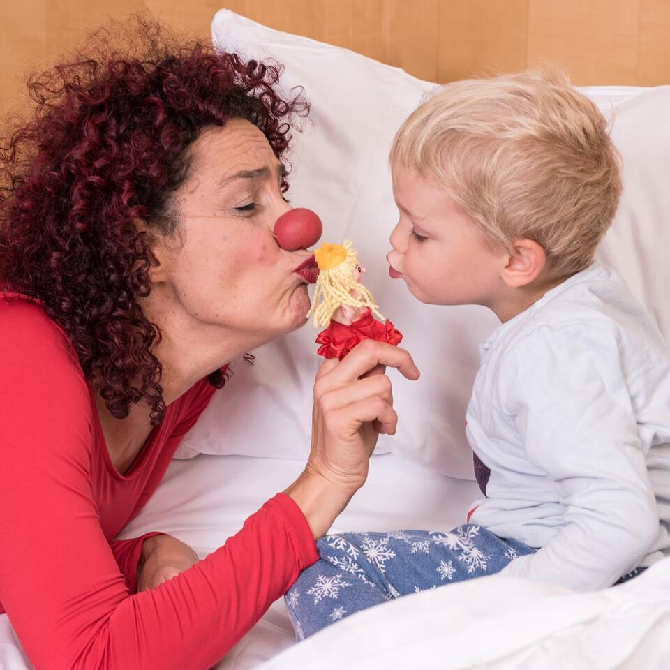 Eine Clownfrau unterhält mit einer kleinen Fingerpuppe ein kleines Kind, das in einem Bett sitzt.