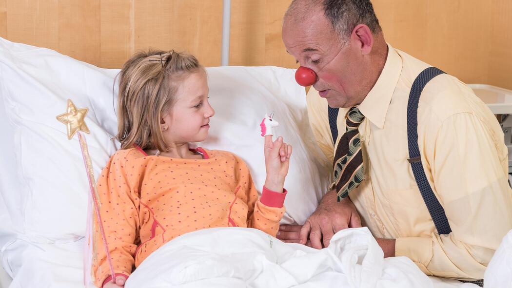 Ein blondes Mädchen liegt in einem Bett. In einer Hand hält sie einen Stab mit einem Stern, auf dem Zeigefinger der anderen Hand sitzt eine Einhorn-Fingerpuppe. Vor ihr schaut ein Clown mit großen Augen auf die Einhornpuppe.
