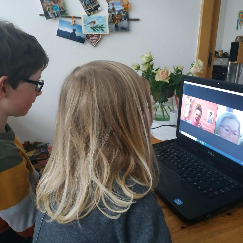 Zwei Kinder sitzen vor einem Laptop und unterhalten sich mit einer Clownin mit roter Nase.